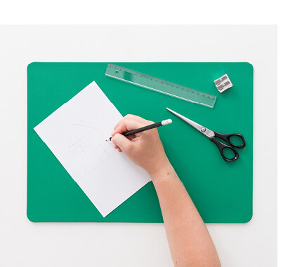 Eine Hand schreibt mit einem Stift auf einem weißen Blatt Papier. Darunter liegt eine quadratische, flache Unterlage aus Kunststoff. Eine Schere, ein Lineal und ein Anspitzer liegen ebenfalls auf der Unterlage.