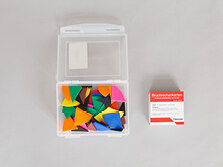 Eine durchsichtige Plaste-Box mit Verschluss, die Kunststoff-Legematerial enthält, das die Bruche von 1/1 bis 1/12 darstellt. Daneben liegen Bruchrechenkarten mit Aufgabenstellungen.