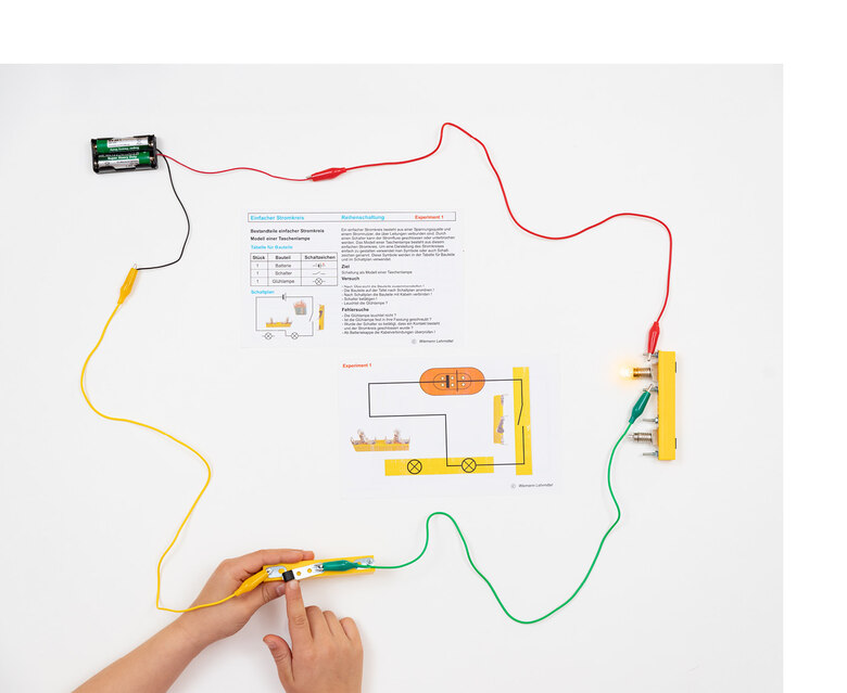 Die bunten Kabel und einzelne Bauteile sind miteinander verbunden. In der Mitte liegt eine Beschreibung des aufgebauten Experiments.