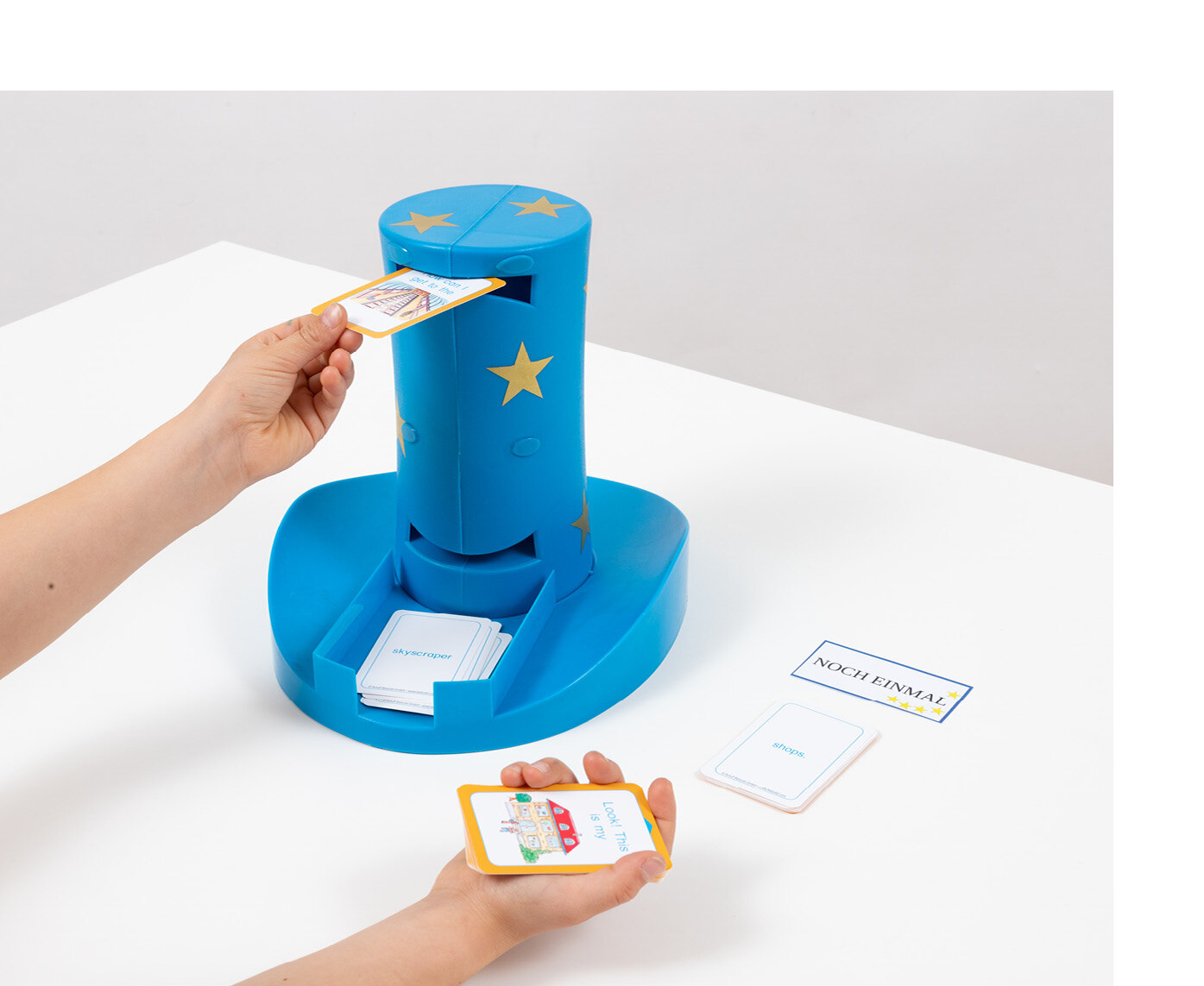 Ein blauer Plastik-Zylinder mit einem Schlitz am oberen und unteren Ende des Zylinders, steht auf einem Tisch. Eine Person steckt mit der linken Hand eine bunte Spielkarte in den oberen Schlitz. In der rechten Hand hält die Person weitere Spielkarten. 