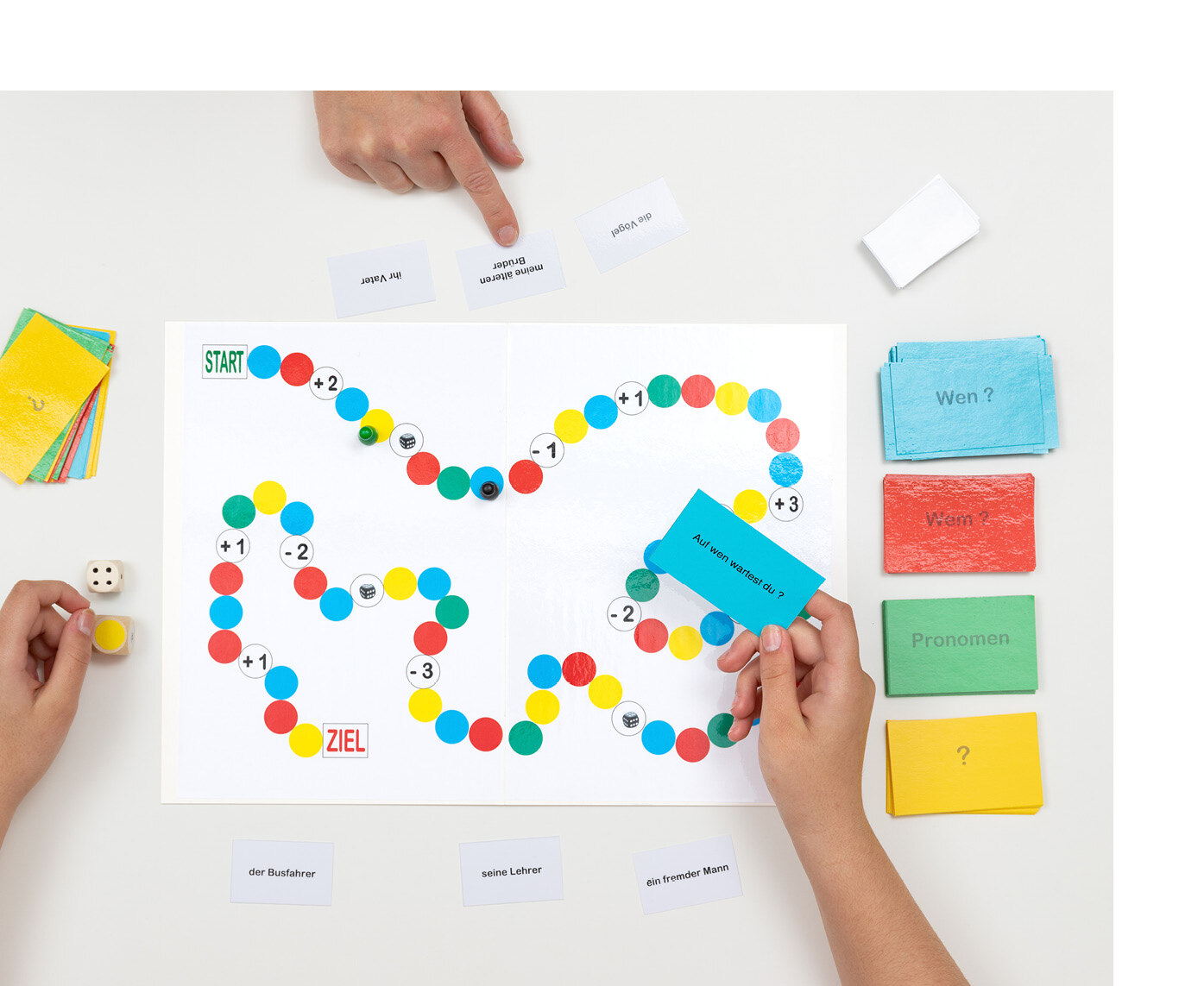 Spielfiguren stehen auf dem Spielfeld und Hände halten farbige Karten in der Hand, auf denen kurze Sätze stehen.